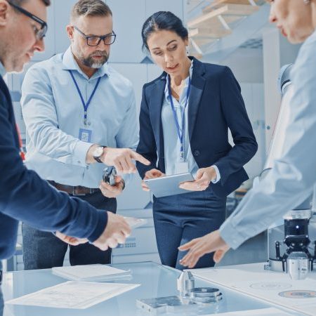 Diverse Team of Industrial Robotics Engineers Gathered Around Table With Robot Arm, They Use Tablet Computer to Manipulate and Program it to Pick Up and Move Metal Component. Bright Facility